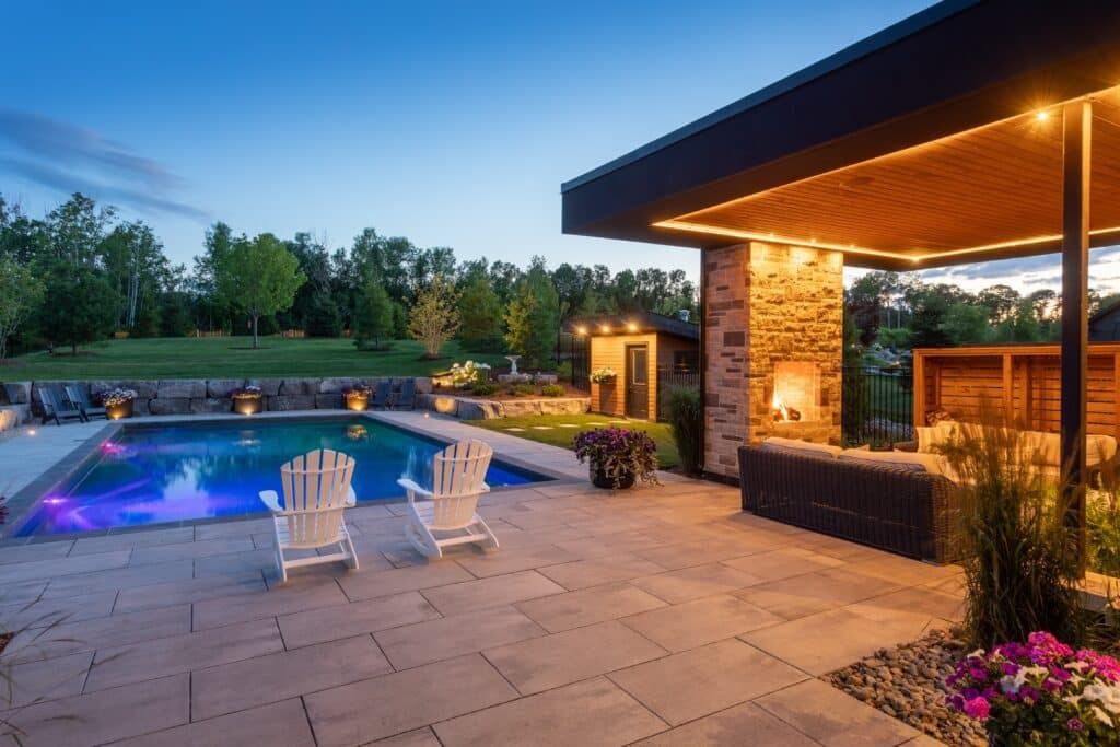 Modern backyard pool with lounge chairs, fireplace patio, and garden lighting at dusk.